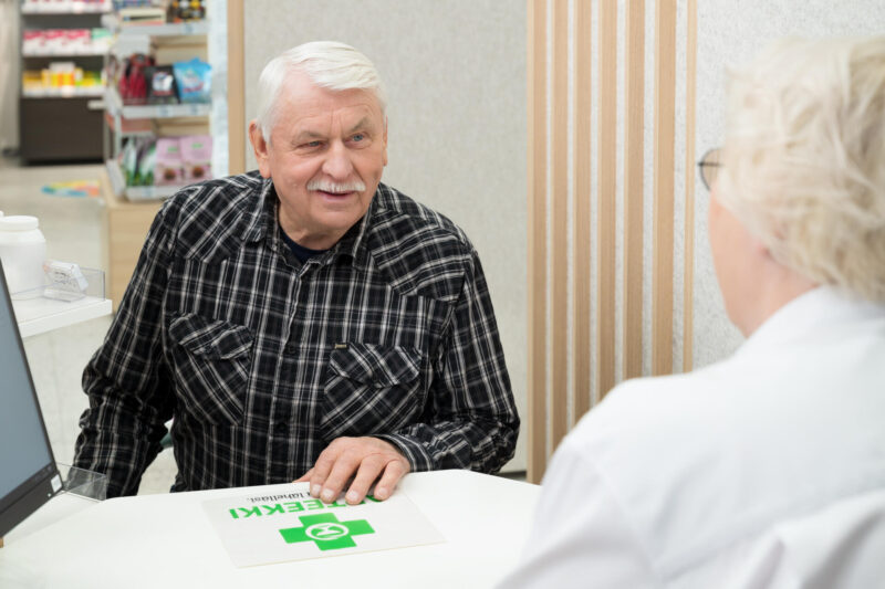 Iäkäs mies apteekin reseptitiskissä juttelemassa farmaseutin kanssa.